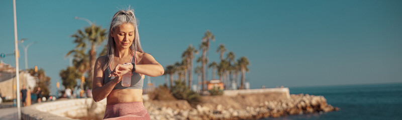 Strong lady in sports clothes sitting by the seashore, holding bottle of water and looking at the watch