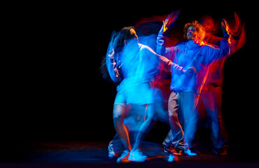 Fototapeta premium Young man and woman dancing hip-hop in sportive style clothes on dark background at dance hall in mixed neon light. Youth culture, hip-hop, movement