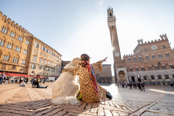 Fototapeta premium Woman sitting with her dog on main square of Siena city with a town hall on background. View from the backside. Concept of travel on tuscany and friendship with pet