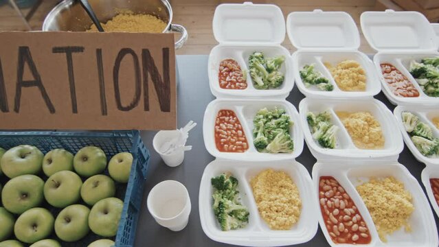 High angle tracking of table with fresh bread, green apples, healthy balanced meals in disposable lunch boxes and handmade cardboard sign with donation inscription indoors at daytime