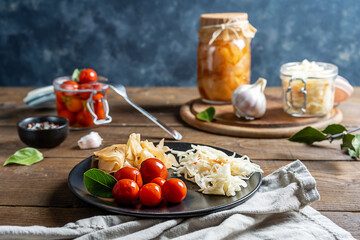 Pickled tomatoes, kimchi, cabbage in jar on cutting board and plate with napkin and fork on wooden table with grey background
