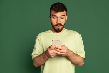 Portrait of young emotive man looking at phone with shocked expression isolated over green studio...