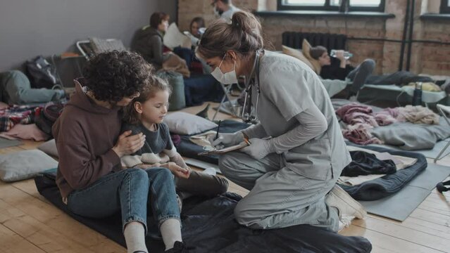 Full shot of female Biracial nurse in scrub suit and face mask checking on Caucasian woman with little son in refugee shelter at daytime