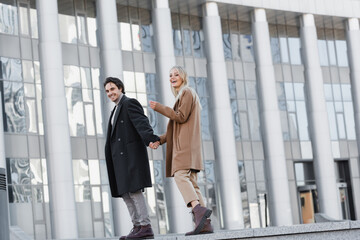 full length of cheerful couple in leather boots and autumn coats holding hands near blurred building.