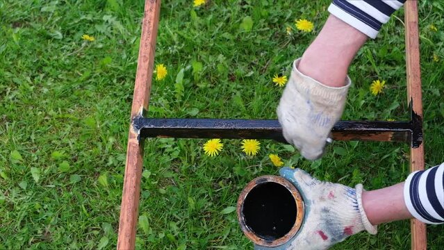 Hands in construction gloves and a vest dip a brush into a jar of black paint and paint an iron staircase, outdoors, against a background of green grass and dandelions.