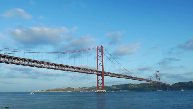 Ponte 25 de Abril, famous suspension bridge at Lisbon, time lapse shot at evening hour. Light clouds quickly fly on sky, small vessels move on waters of Tagus river