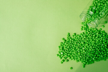 GRANULES OF GREEN WAX LIE ON A GREEN BACKGROUND, POURED FROM A TRANSPARENT JAR. SPACE FOR TEXT