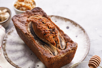 Chocolate banana bread with walnuts on a white plate with brown rim and ingredients on a grey neutral background with creative lighting, top view