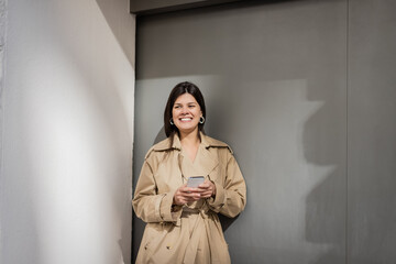 happy woman in trench coat texting on smartphone near wall.