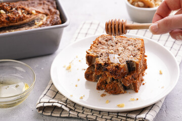 pouring honey onto Slices of Chocolate banana bread with walnuts on a checkered kitchen napkin and ingredients on a grey neutral background