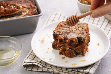pouring honey onto Slices of Chocolate banana bread with walnuts on a checkered kitchen napkin and ingredients on a grey neutral background