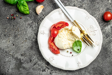 Salad with creamy Italian Burrata Cheese Served with Olive Oil, Basil Leaves and cherry tomatoes on white plate on a dark background. recipe top view