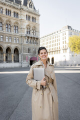 Fototapeta premium happy freelancer in beige trench coat walking with laptop on street of vienna.