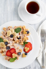 Belgian waffles with fruit and mint leaves on a white plate, cutlery and a cup of tea.