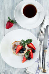 On a gray table is a blurred image of a plate of pancakes and strawberries, cutlery and a cup of coffee.