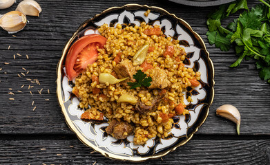 Eastern Uzbek cuisine. Pilaf from Bulgur groats with veal and lamb with carrots. Bulgur pilaf lies in an Uzbek plate with a national pattern on a woody black background.