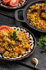 Eastern Uzbek cuisine. Pilaf from Bulgur groats with veal and lamb with carrots. Bulgur pilaf lies in an Uzbek plate with a national pattern on a woody black background.