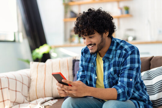Young joyful Asian man using mobile phone and smiling sitting at home on the couch. Male holds smartphone in his hands looking at the screen, reads a message, plays games