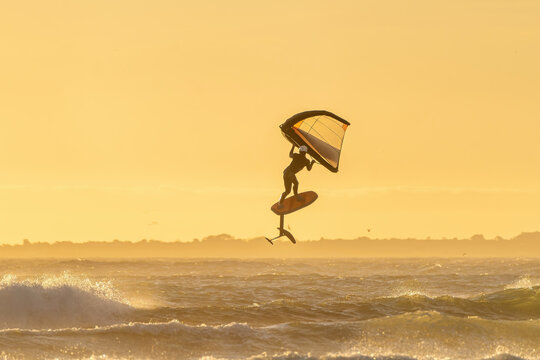 Focus on jump with wing foil equipment over a big ocean wave during sunset in Cape Town. Wing Foiling the new trend sport. Golden Background horizontal image with windy ocean and waves. 