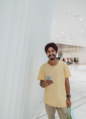 Portrait Of A Handsome Sikh Man Using Mobile Phone And Holding Shopping Bags While Standing In The...