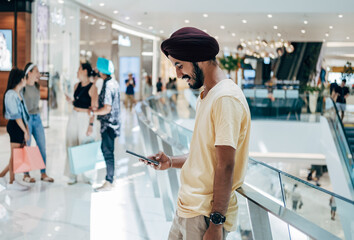 Happy Man Using Mobile Phone At The Shopping Mall. 
Cheerful smiling Indian man with turban typing text message on his smartphone while standing at the modern shopping mall. - obrazy, fototapety, plakaty