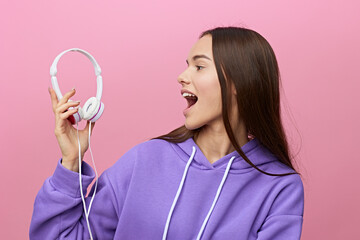 a pretty, happy woman stands on a pink background in a purple hoodie and holding headphones in her...