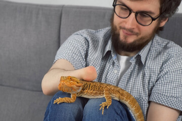 Disabled man with amputated two stump hands with animal bearded agama sitting on sofa at home and stroking him, lovely pet friend. Animal therapy. Problem of adaptation life people with disabilities. - obrazy, fototapety, plakaty
