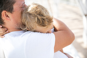 Fototapeta premium On a warm summer day, a Caucasian man gently and lovingly hugs his child whose head lies on his strong shoulder. Depersonalization.
