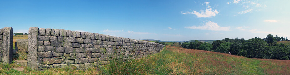 panoramic rural summer scene with a gate in a long stone wall surrounded by meadow grass with tree and west yorkshire dales hills in the distance