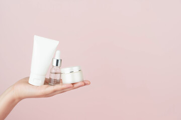 Skincare routine​ for healthy skin. Woman hand holding facial foam tube, serum bottle, moisturizing​ cream on pink background. Beauty​ and cosmetic concept. - obrazy, fototapety, plakaty
