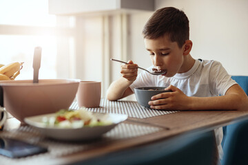 Caucasian small teen boy eating cereals with milk and drinking tea at table in morning.