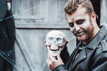 Scary man with dreadlocks looking at skull. Celebrating halloween. Terrifying black white face monster makeup and stylish costume, image.Horror,fun at children's party in barn on street.Shirt,jackets