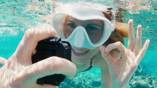 Young woman swims underwater with action camera and shows OK signal with her hand