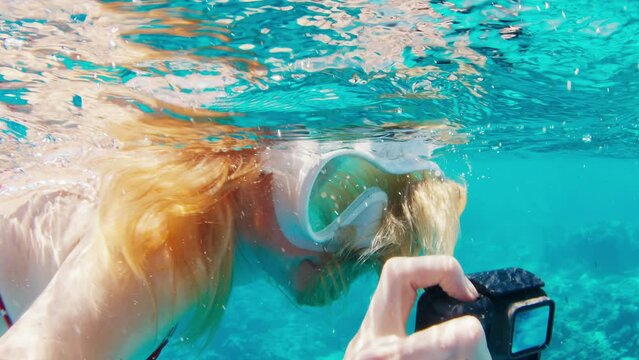 Woman with mask swims underwater with action camera and films a content