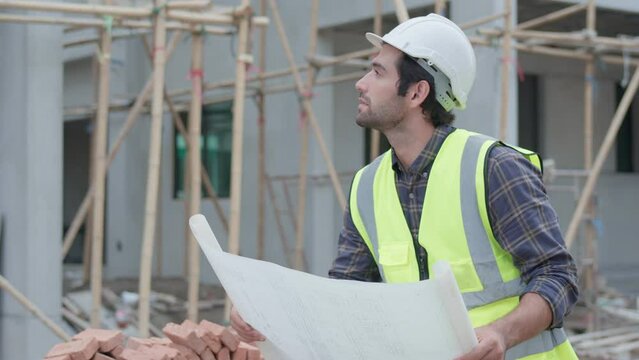 Professional male civil engineer or architect wear safety helmet. While handsome engineer working, holding blueprint plan check look around building project on construction site. Real estate concept.