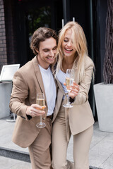 cheerful couple in beige stylish suits holding champagne glasses on street in city.