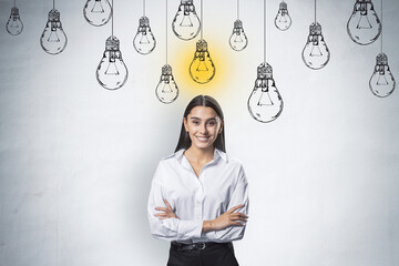 Creative idea and startup concept with happy and smiling young woman in white shirt at light wall background with hand drawn white lightbulbs and yellow one switched on - obrazy, fototapety, plakaty