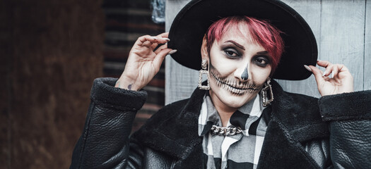 Scary woman, girl celebrating halloween.Terrifying black skull half-face skeleton makeup and witch costume in stylish image. Jacket, hat.Horror,fun at photoshoot, holiday party near barn on street © velirina