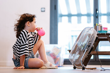 Little girl at home sitting on the floor inflating balloon and playing with fan