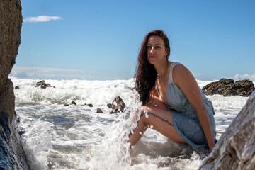 A woman in a blue dress at the beach on a sunny day. The blue sky has some cloud cover. The model sits amongst the rocks as the sea washes waves over her