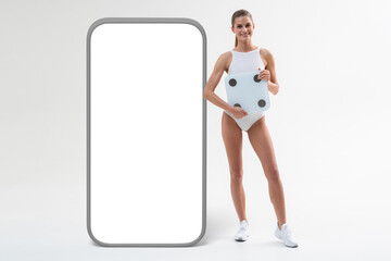 Sporty fitness coach with bathroom scales standing next to huge smartphone mockup