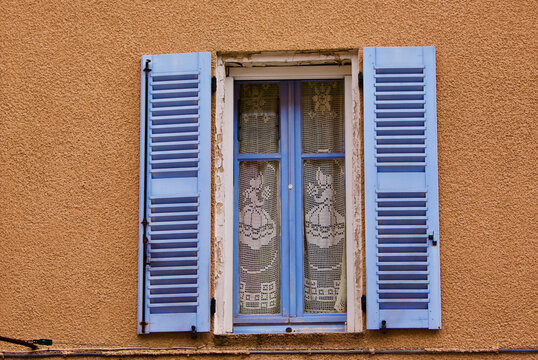 Brown stone building with blue window shutters and window with lace curtain in France.