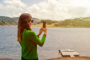 Young woman in front of a lake taking a photo using her cell phone