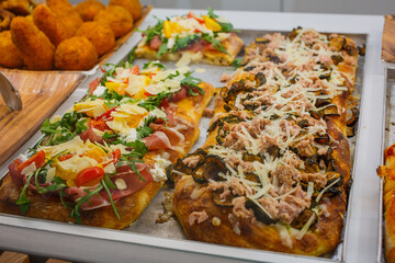 Pizze romane con zucchine fritte, tonno, mozzarella, provole del monaco, stracciata di bufala, pomodori rossi e gialli, rucola e prosciutto crudo in esposizione in una vetrina di una rosticceria