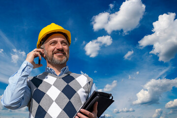 Smiling Engineer Talking On Mobile Phone Against Blue Sky With White Clouds. Happy Manager In Yellow Hardhat Standing And Using Smart Phone Outdoors.