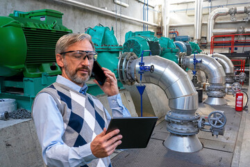 Elegant CEO Looking At Digital Tablet Screen And Talking On Smartphone Beside Large Generators And Pipes. Confident, Gray-Haired Manager or Businessman. Man Using Smartphone And Digital Tablet.