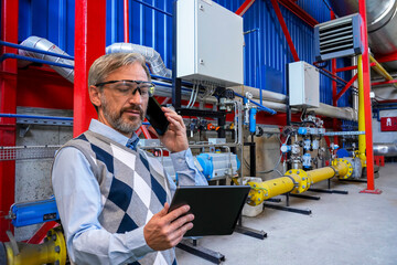 Chief Executive Officer Looking At Digital Tablet Screen And Talking On Smart Phone In Industrial Interior. Confident, Elegant, Gray-Haired CEO or Businessman. Man Using Smartphone And Digital Tablet.