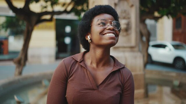 Beautiful girl in a good mood listening music and dancing outdoors. African American woman looking inspired sitting on the street and listening music in wireless earphones
