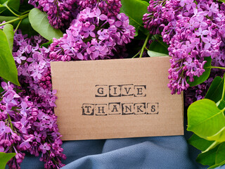A piece of cardboard with words Give Thanks on it next to some lilac flowers and leaves