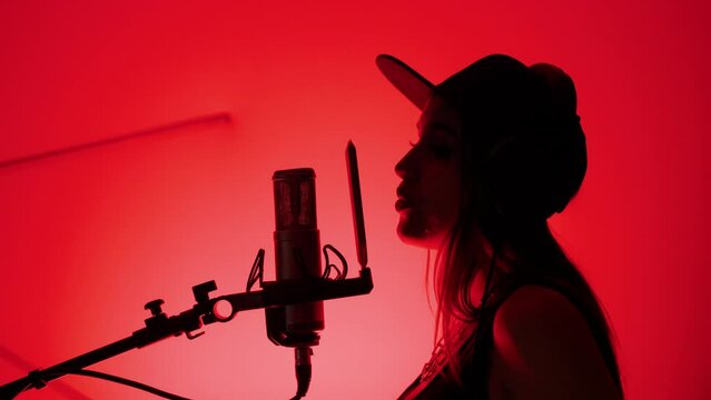 Silhouette of profile woman singer in cap and headphones artist sings song in recording studio. Red Background. Musician stands near studio microphone. Music production studio concept
